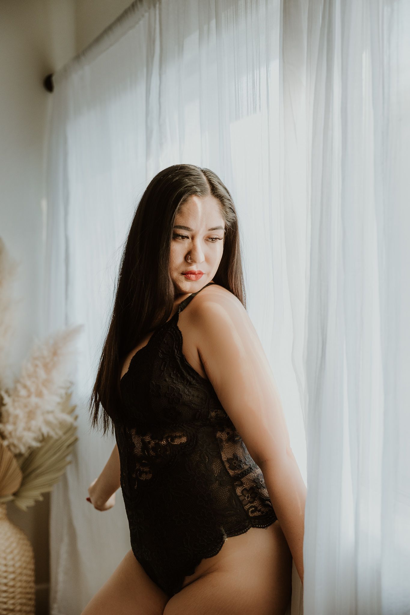 Intimate boudoir photo with black lace bodysuit against white curtain backdrop.