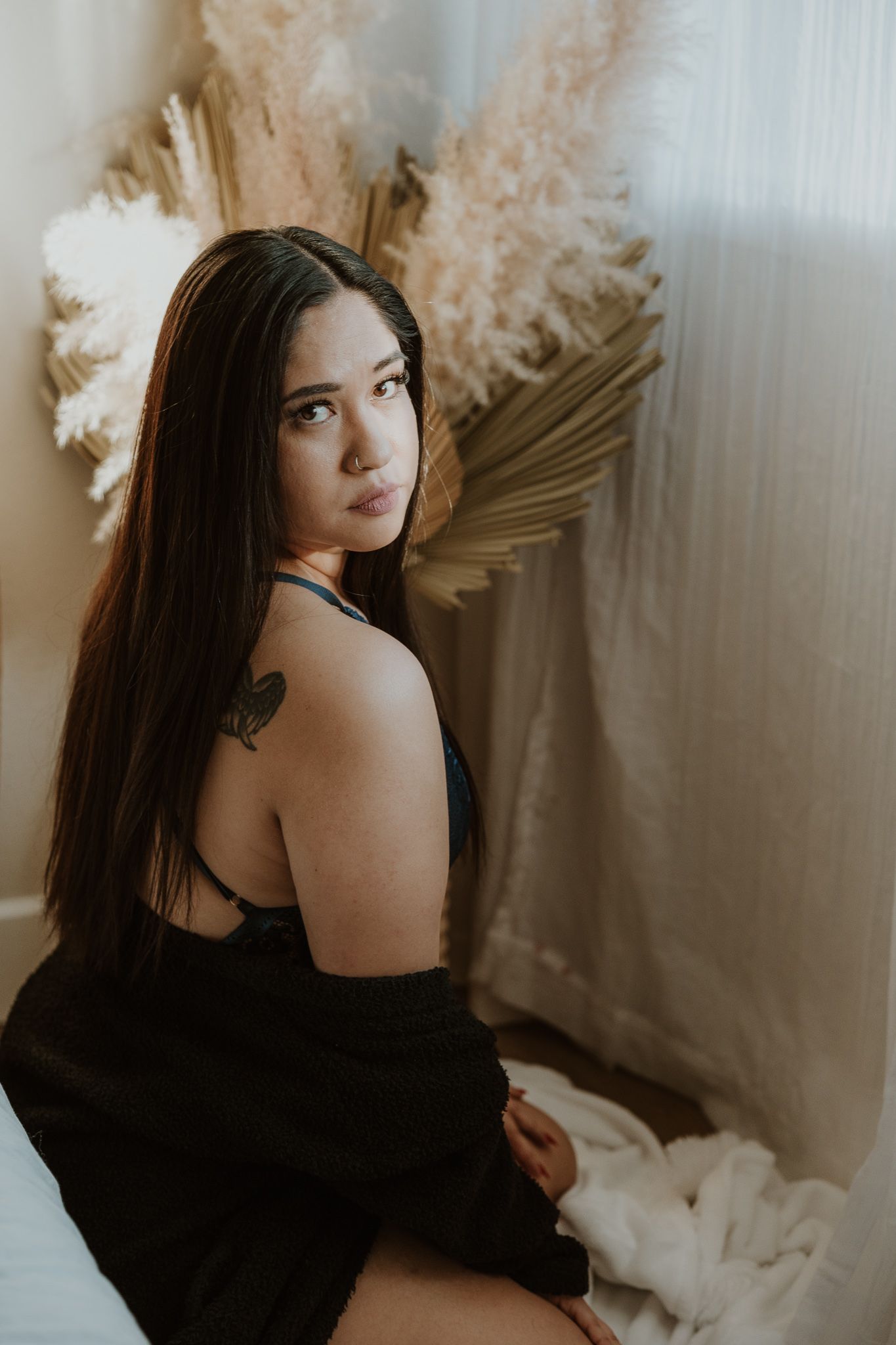 Moody boudoir portrait with long dark hair against white curtains and dried floral decor.