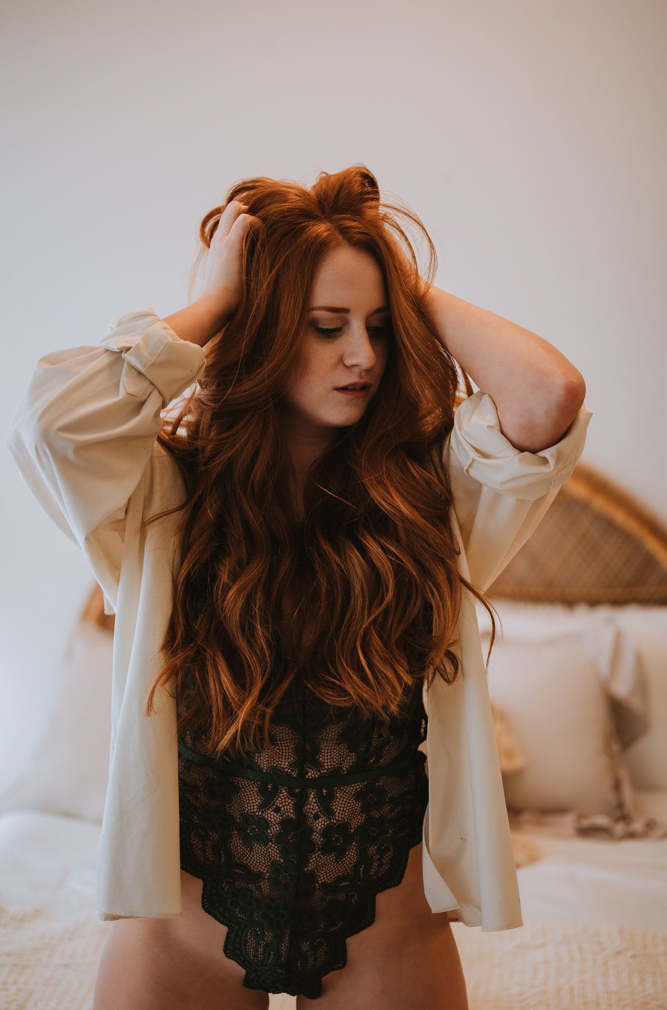 Woman in white shirt and lace bodysuit running hands through long red hair on bed.