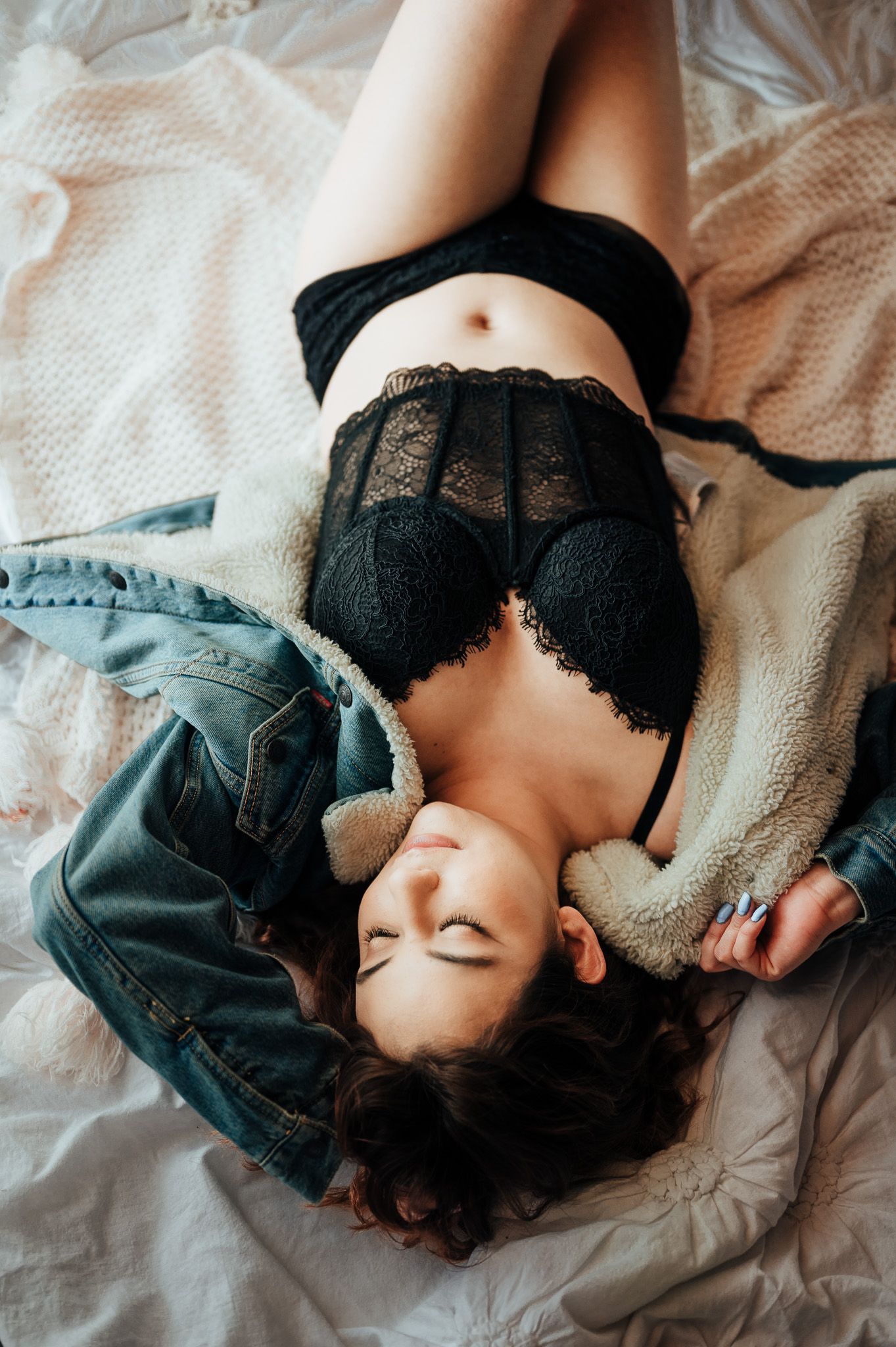 Person in black lace lingerie lying on white bedding in artistic boudoir pose.