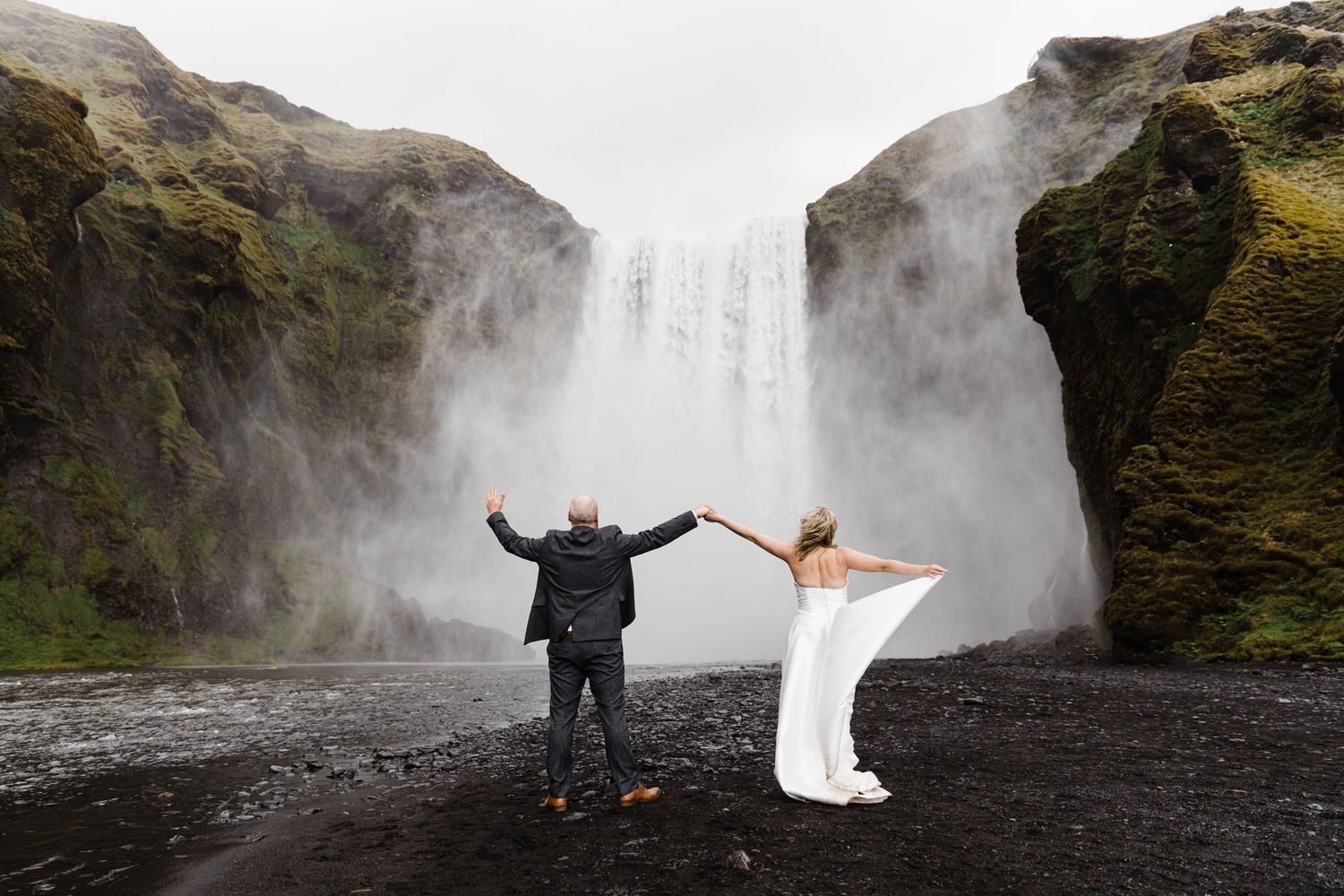 A couple in wedding attire poses dramatically in front of a majestic waterfall in Iceland.