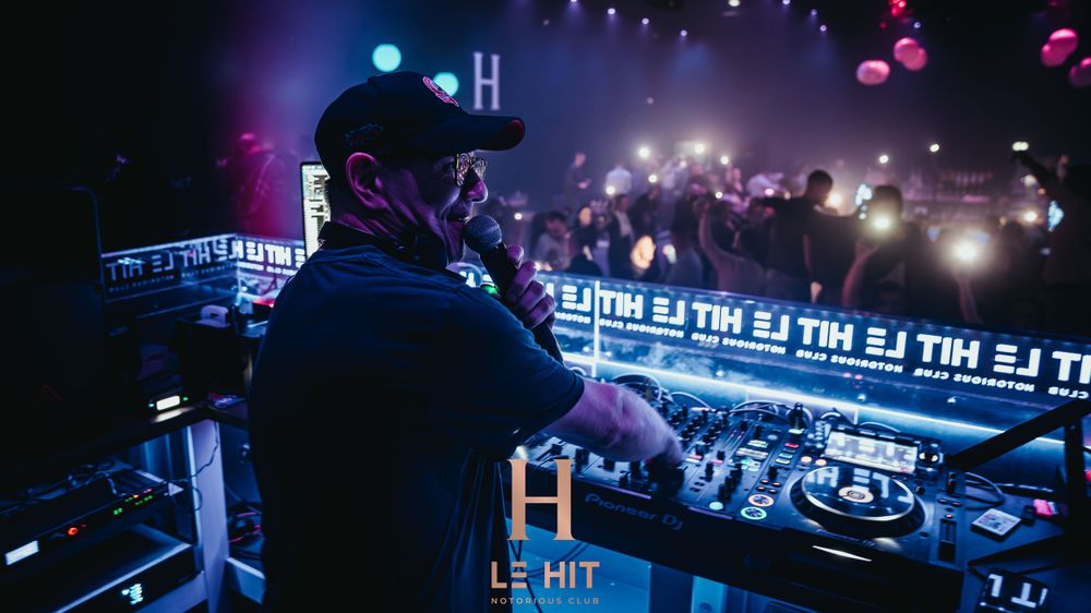 DJ performing at Le Hit nightclub with colorful lights and an energetic crowd behind him.