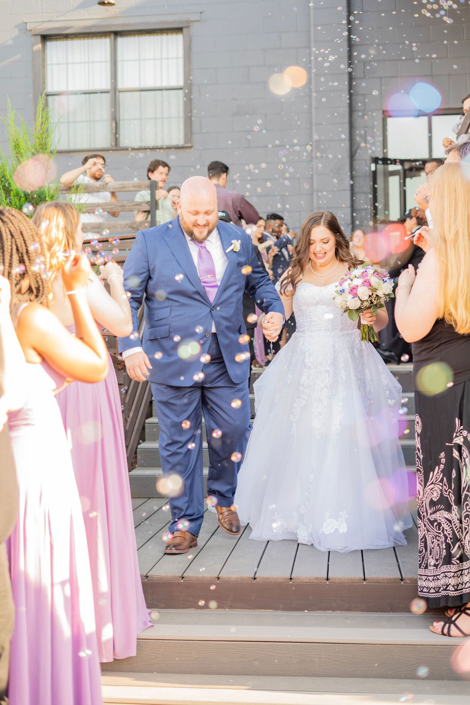 Bride and groom walk through bubble send-off surrounded by guests in lavender dresses outdoors.