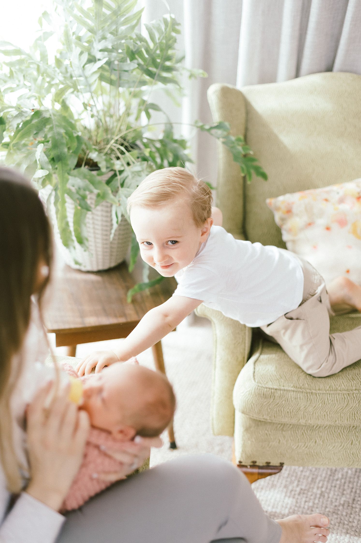 Toddler in white shirt leans over armchair during baby's feeding time.