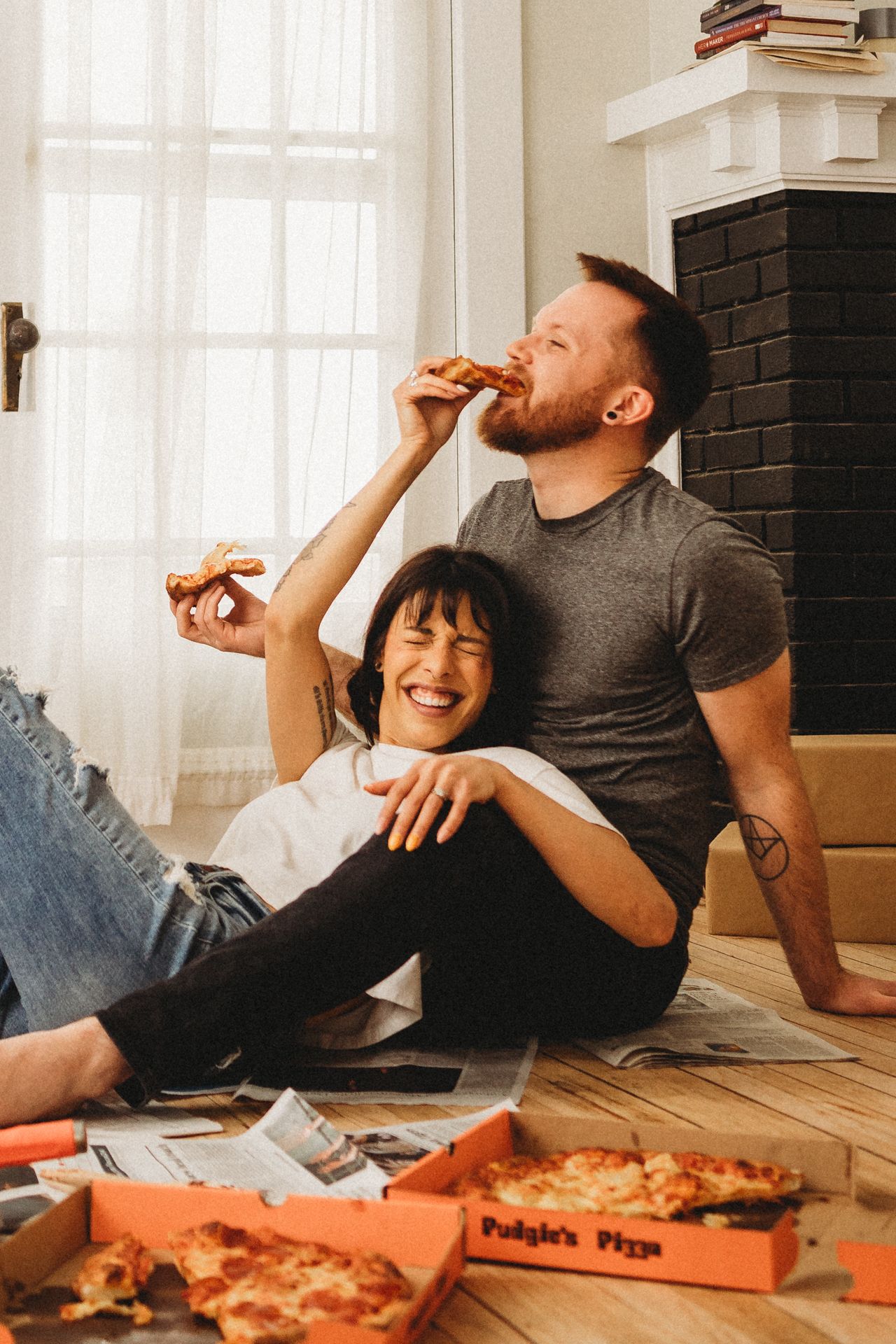 Couple enjoying pizza while sitting on floor near fireplace with delivery boxes.
