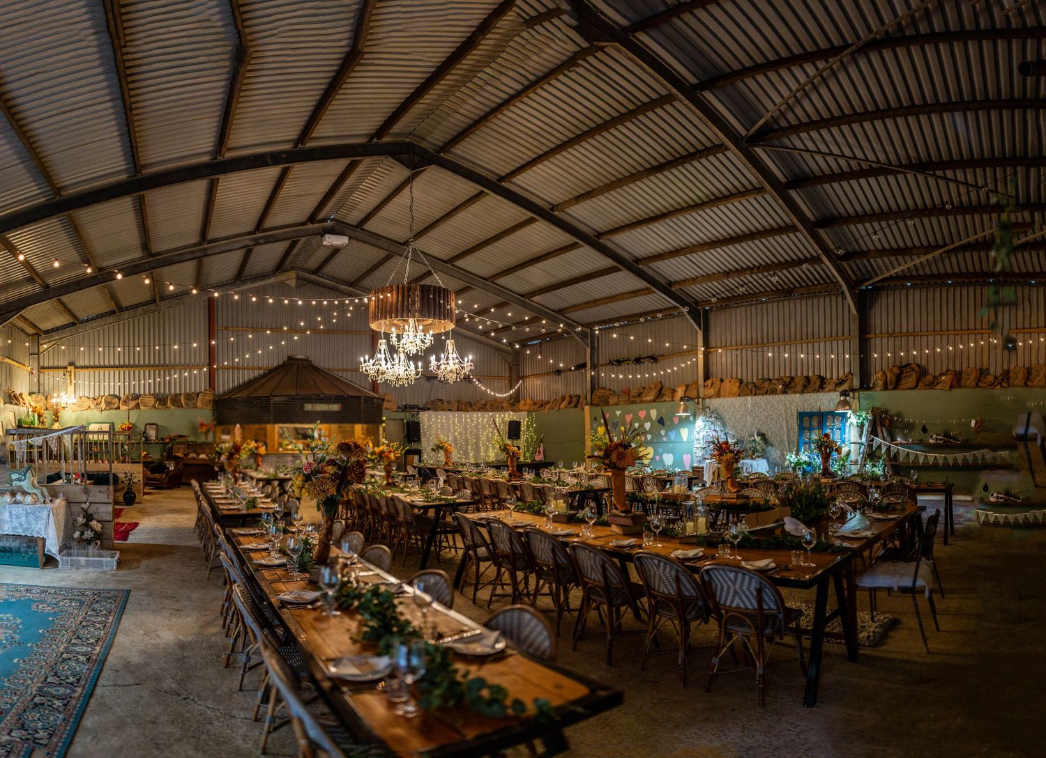 Wide view of barn wedding reception with long tables, chandeliers and string lights creating ambient lighting.