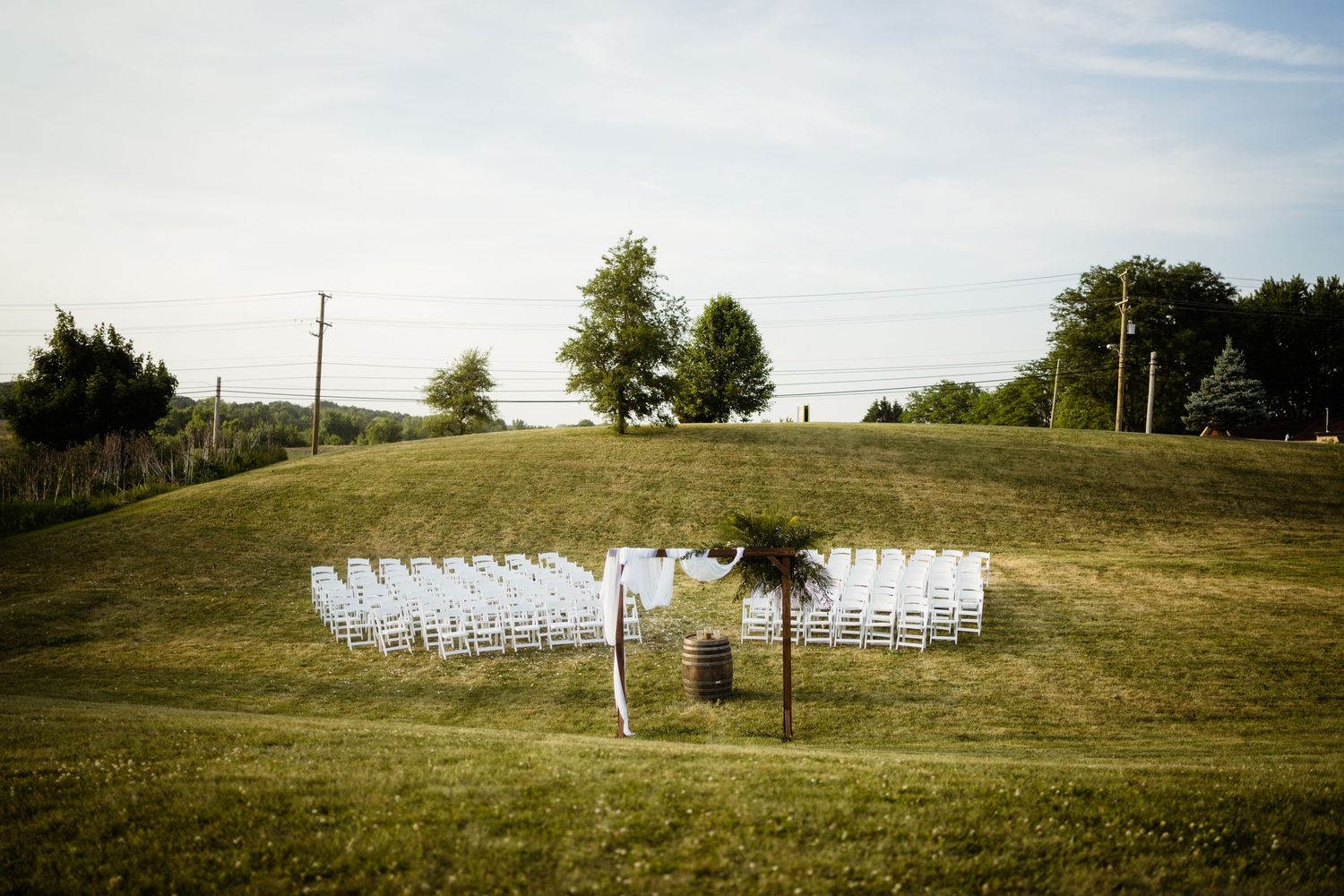 White chairs arranged in rows for an outdoor ceremony on a grassy hilltop with trees in the background.