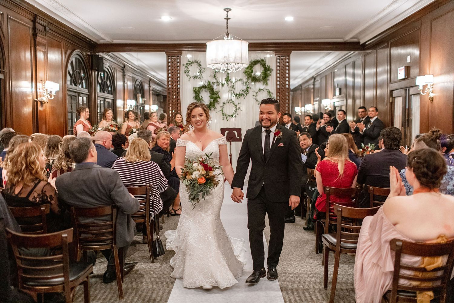 Wedding celebration in an elegant wood-paneled hall with guests seated in rows.