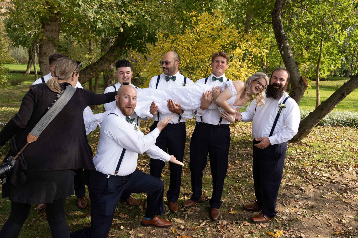 A group of groomsmen pose playfully outdoors in formal attire with white shirts and suspenders.