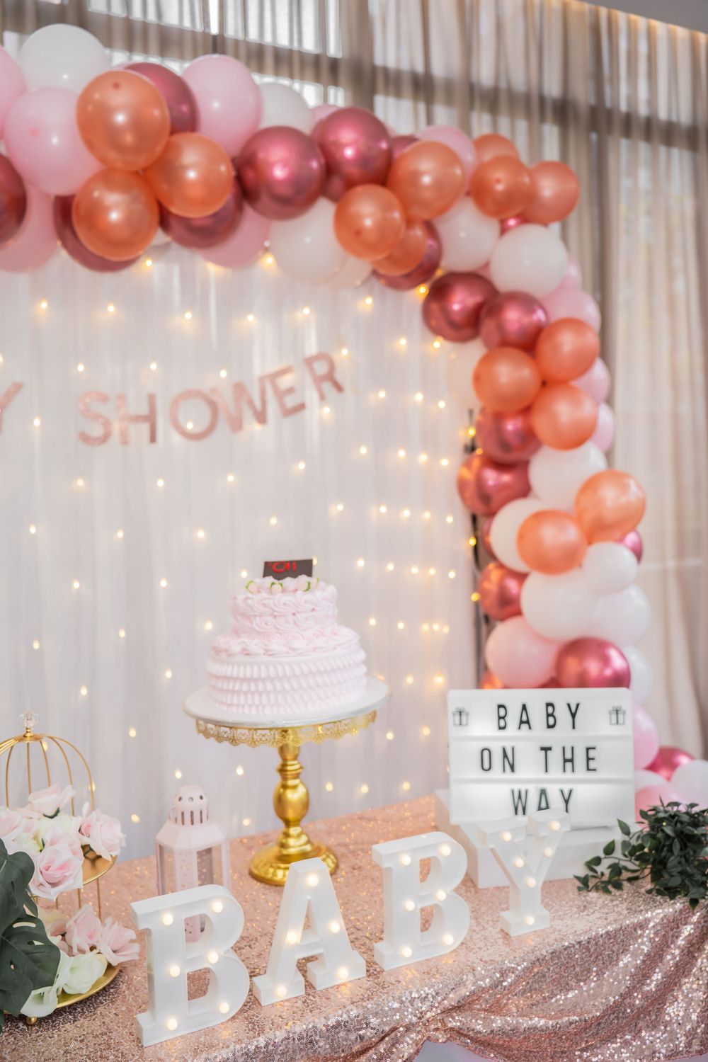 Baby shower decoration with pink and coral balloon arch, cake, and light-up signage.