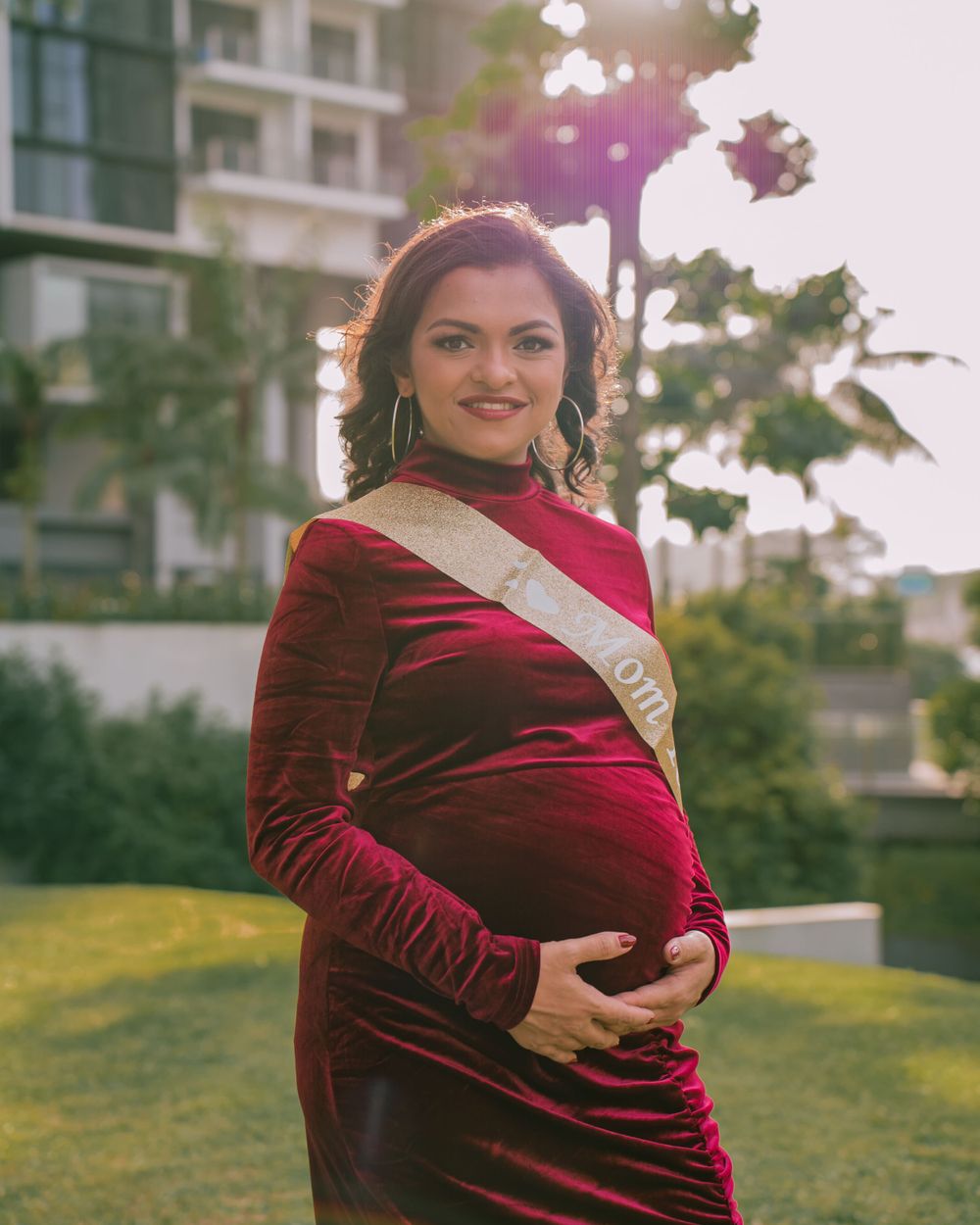 Expectant mother in a burgundy velvet dress poses outdoors at sunset.