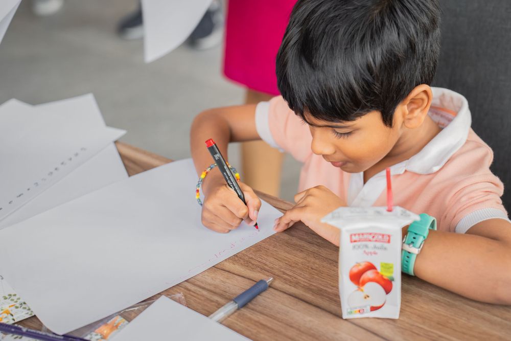 Child doing homework while drinking from a juice box at desk.