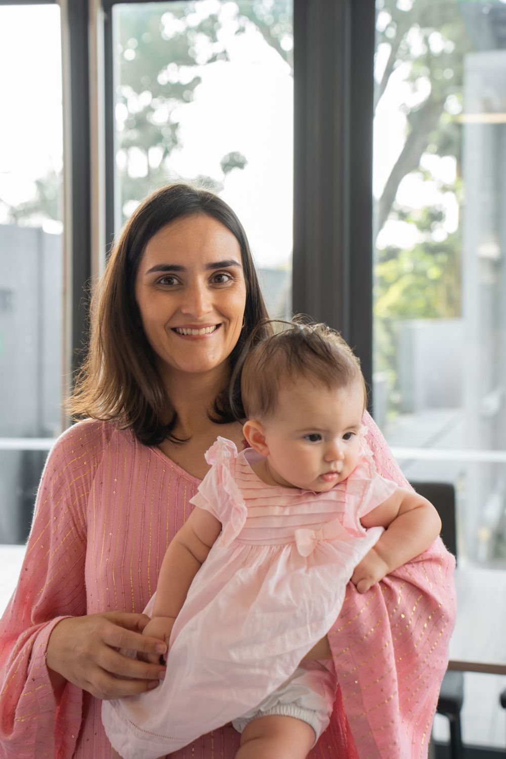 A parent and baby in matching pink outfits share a tender moment by a bright window.