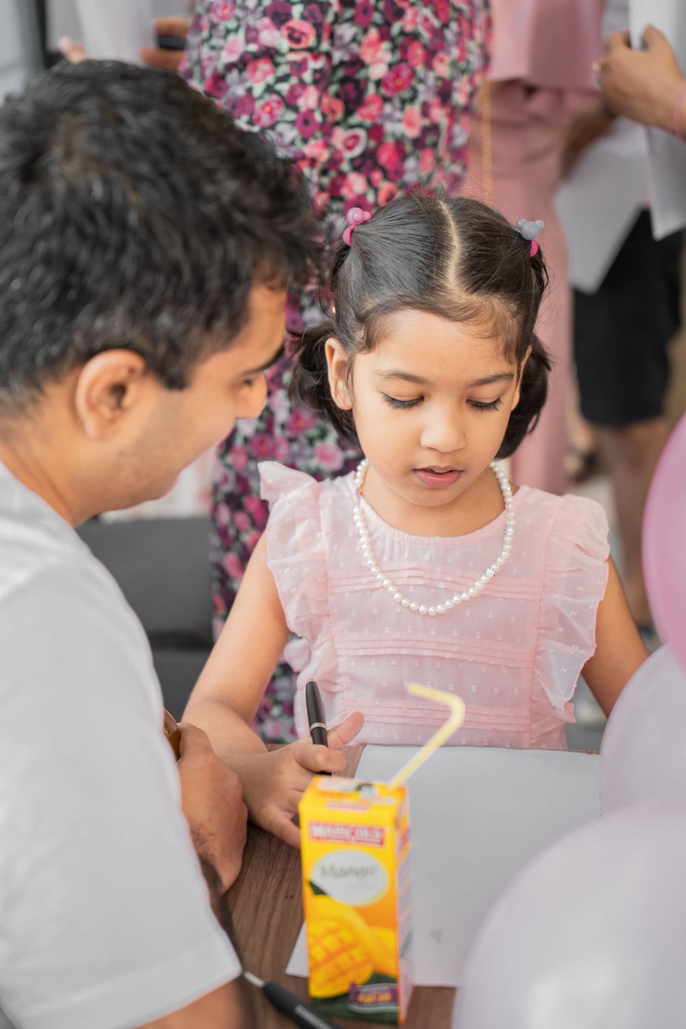 Child in pink dress with pearl necklace drinking from juice box.