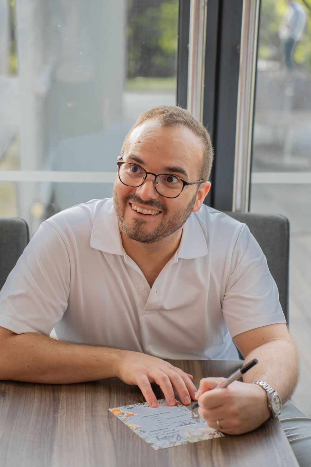 Person in white polo shirt smiles while writing at a wooden desk.