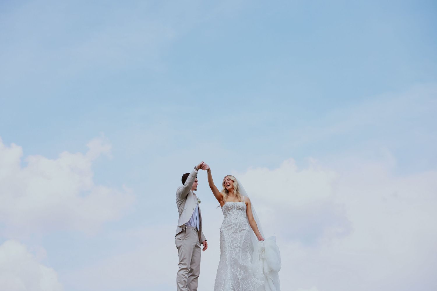 Couple in wedding attire pose against bright blue sky background.