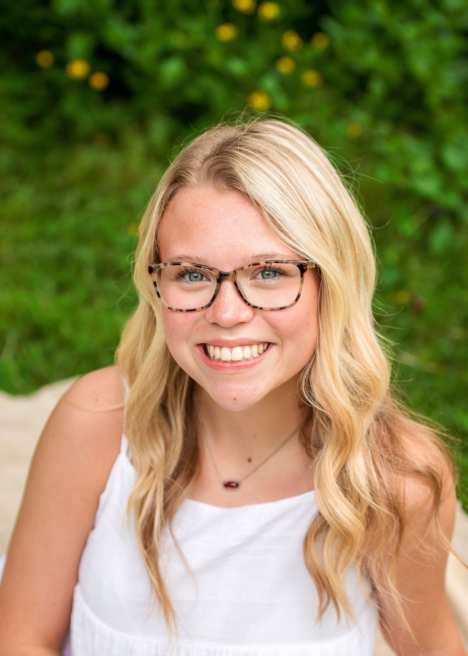 Portrait of a young woman with blonde wavy hair and glasses smiling outdoors against a green background.