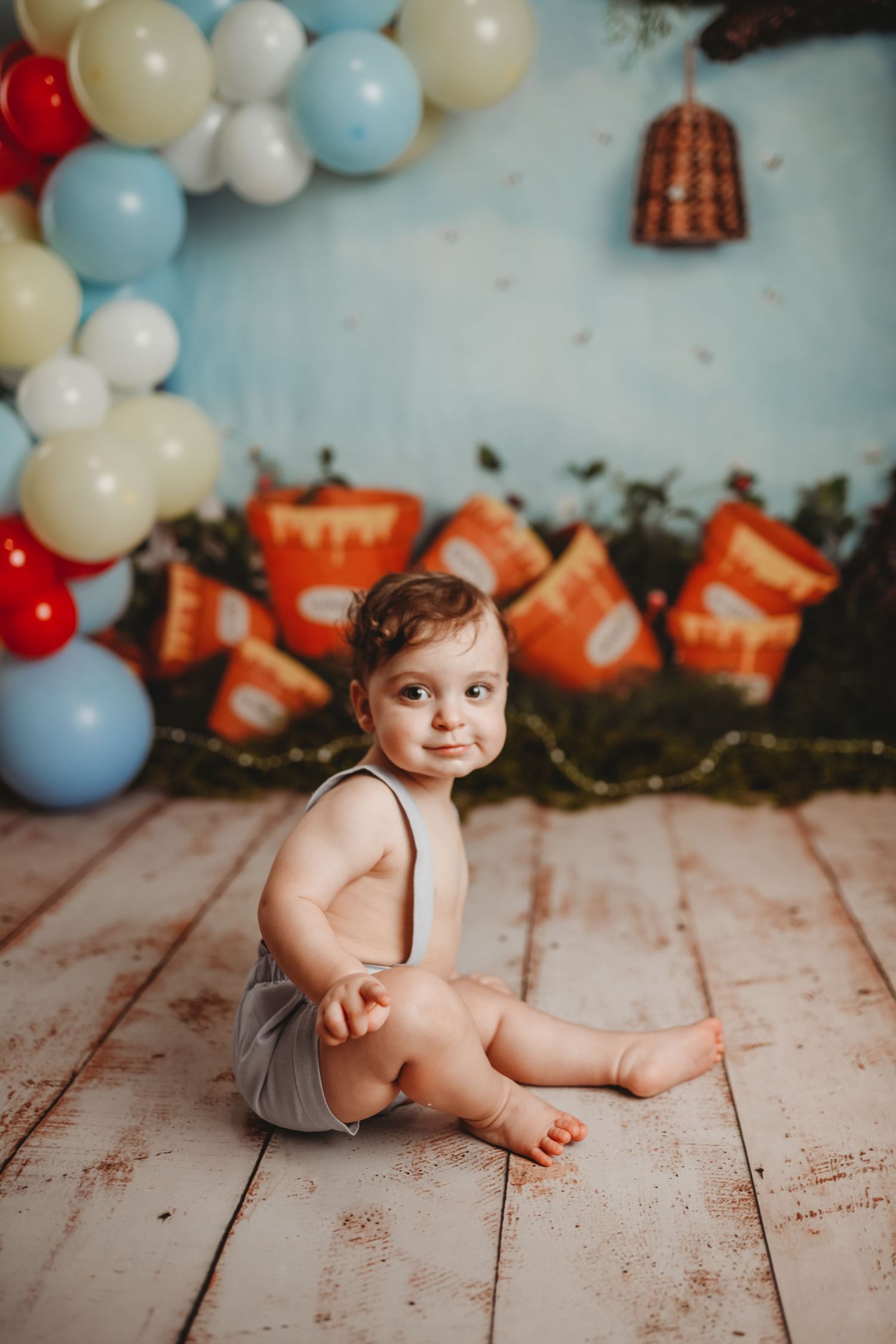 Baby in gray outfit sits on rustic floor with blue and white balloons and orange decorations in background.