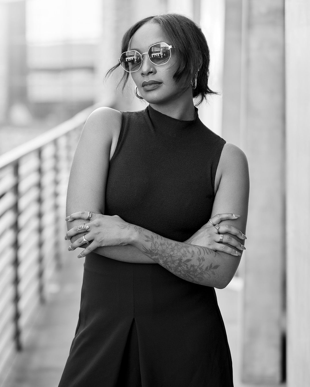 Black and white portrait in sleeveless turtleneck showing tattooed arms and sunglasses.