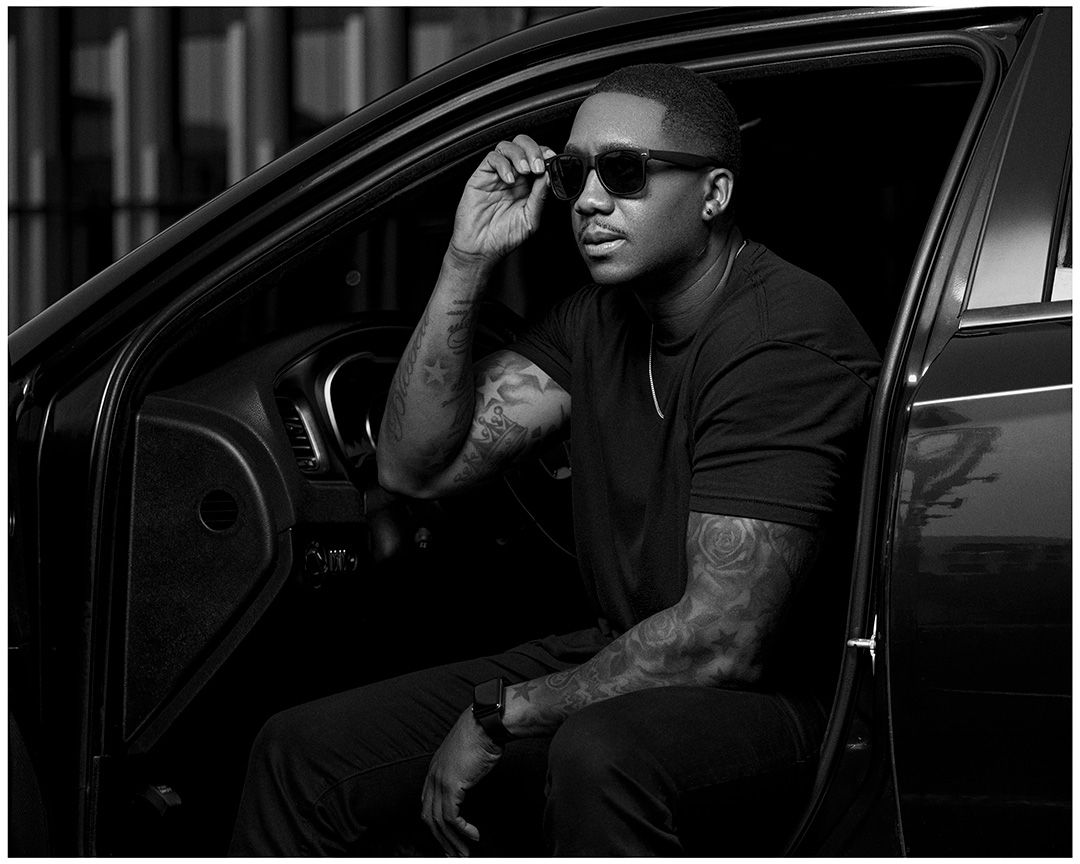 Monochrome image of someone sitting in a car wearing sunglasses and adjusting their hat.