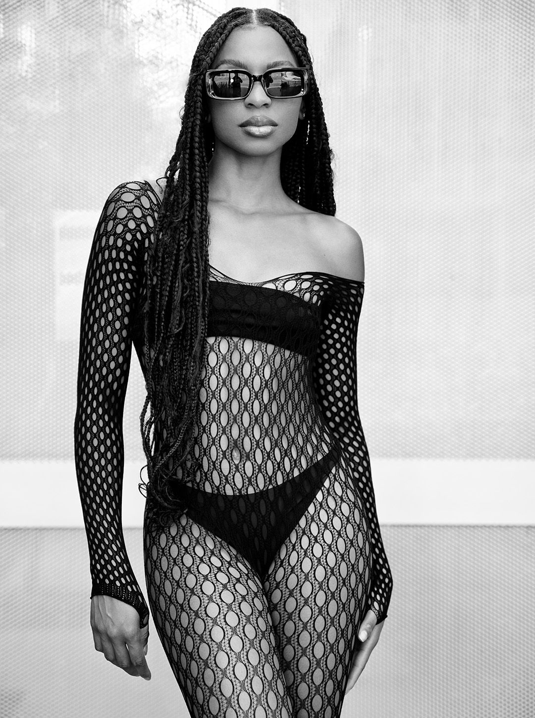 Fashion model wearing a black fishnet bodysuit and sunglasses poses in black and white photo.