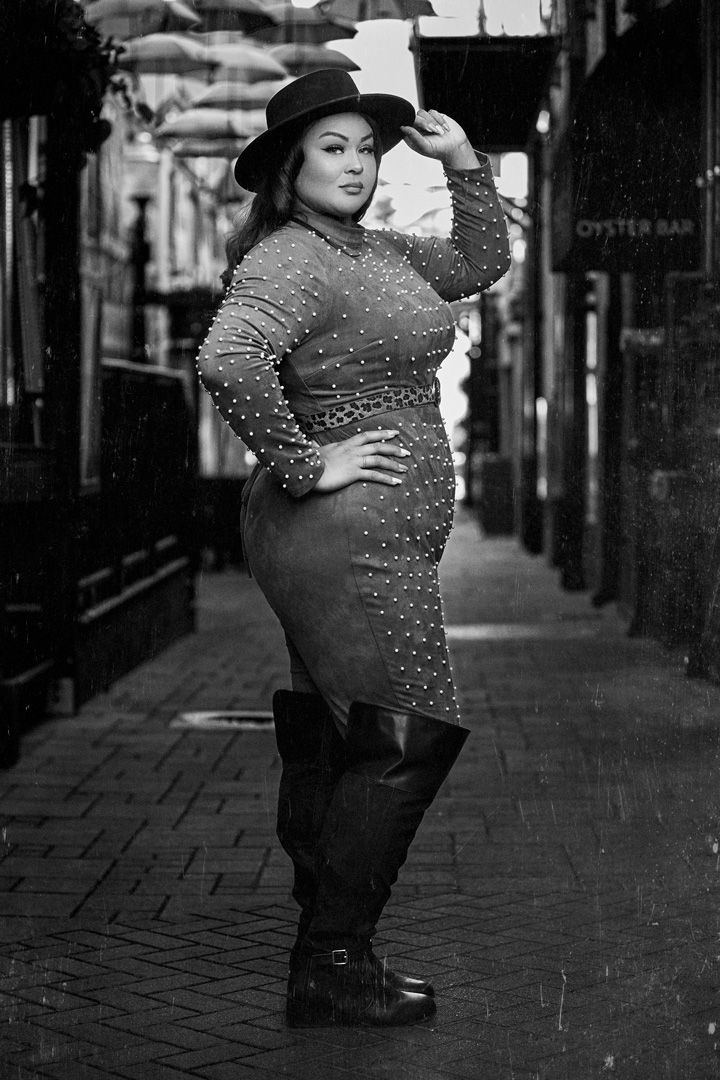 Glamorous black and white photo of a curvy model in a sequined dress and hat on a city street.
