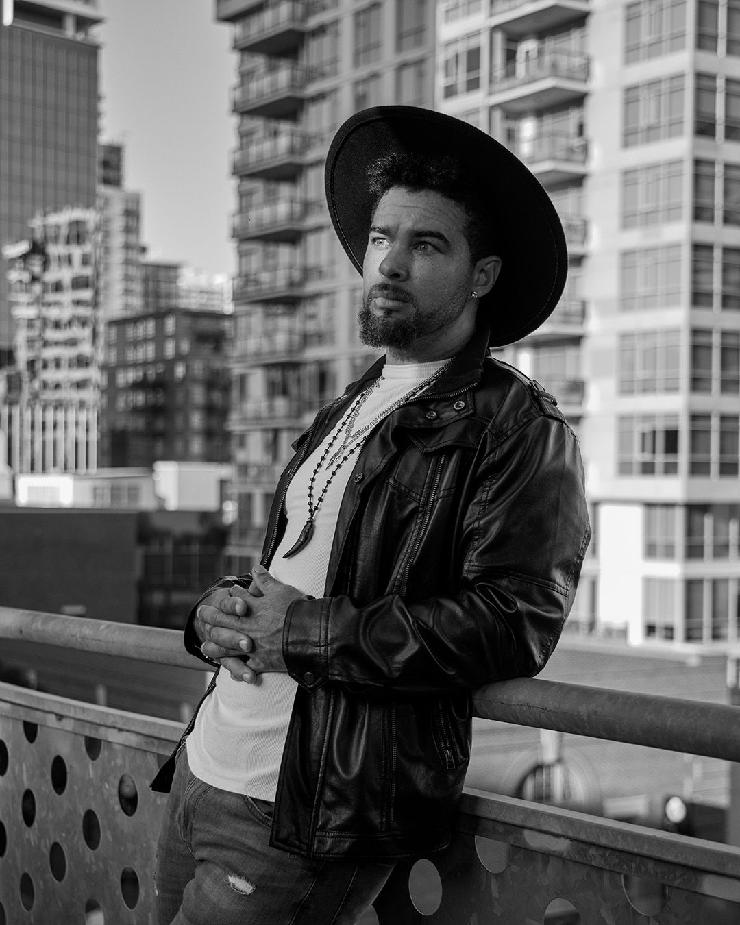 Stylish black and white portrait of someone wearing a wide-brimmed hat and leather jacket against urban backdrop.