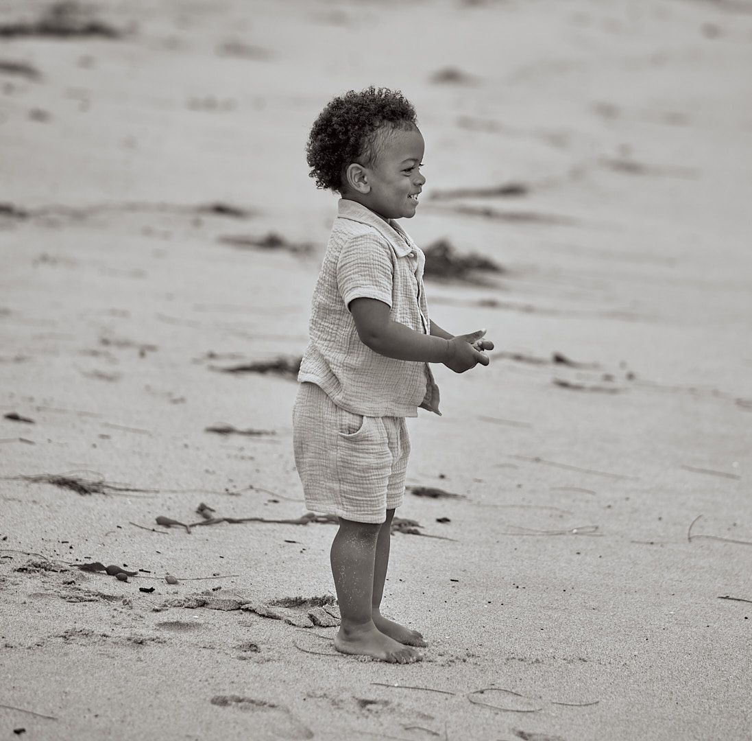 Black and white photo of toddler playing on sandy beach wearing casual summer outfit.