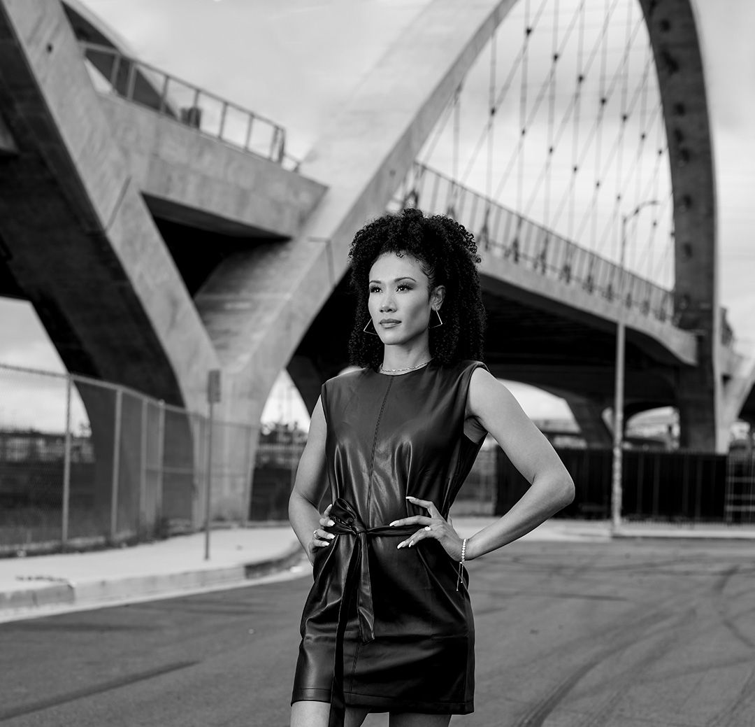 Urban fashion portrait of someone in a black dress posing under architectural bridge structure.