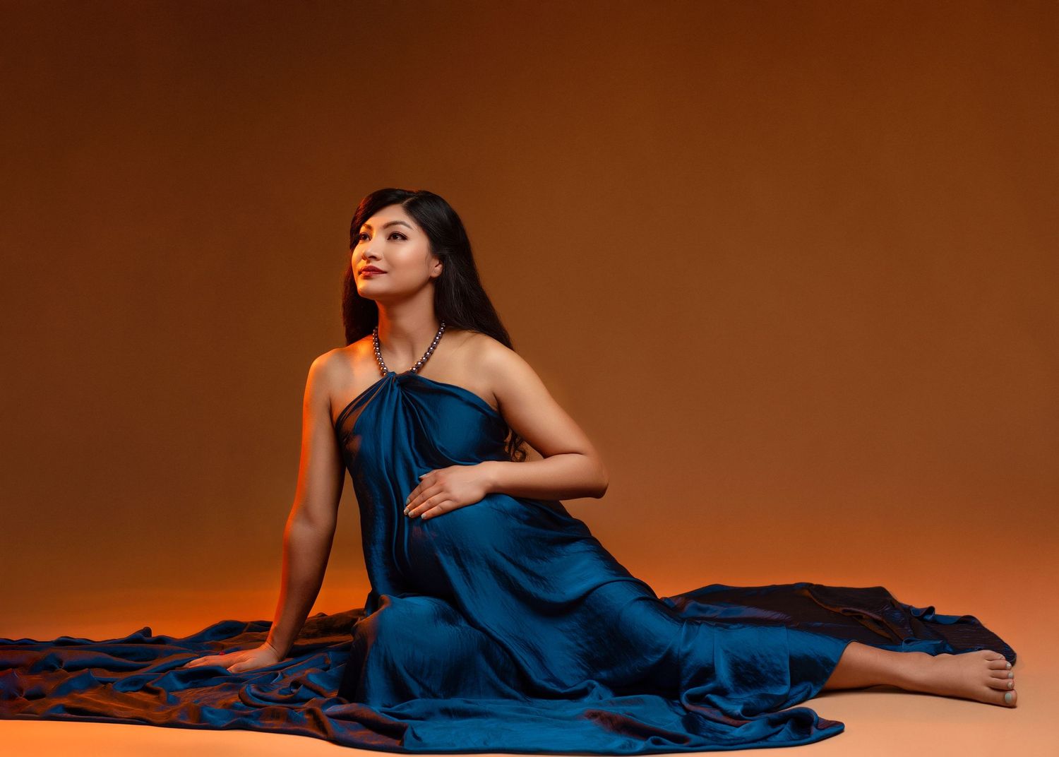 an expectant mother lifts her face while laying on the ground in a flowy blue dress