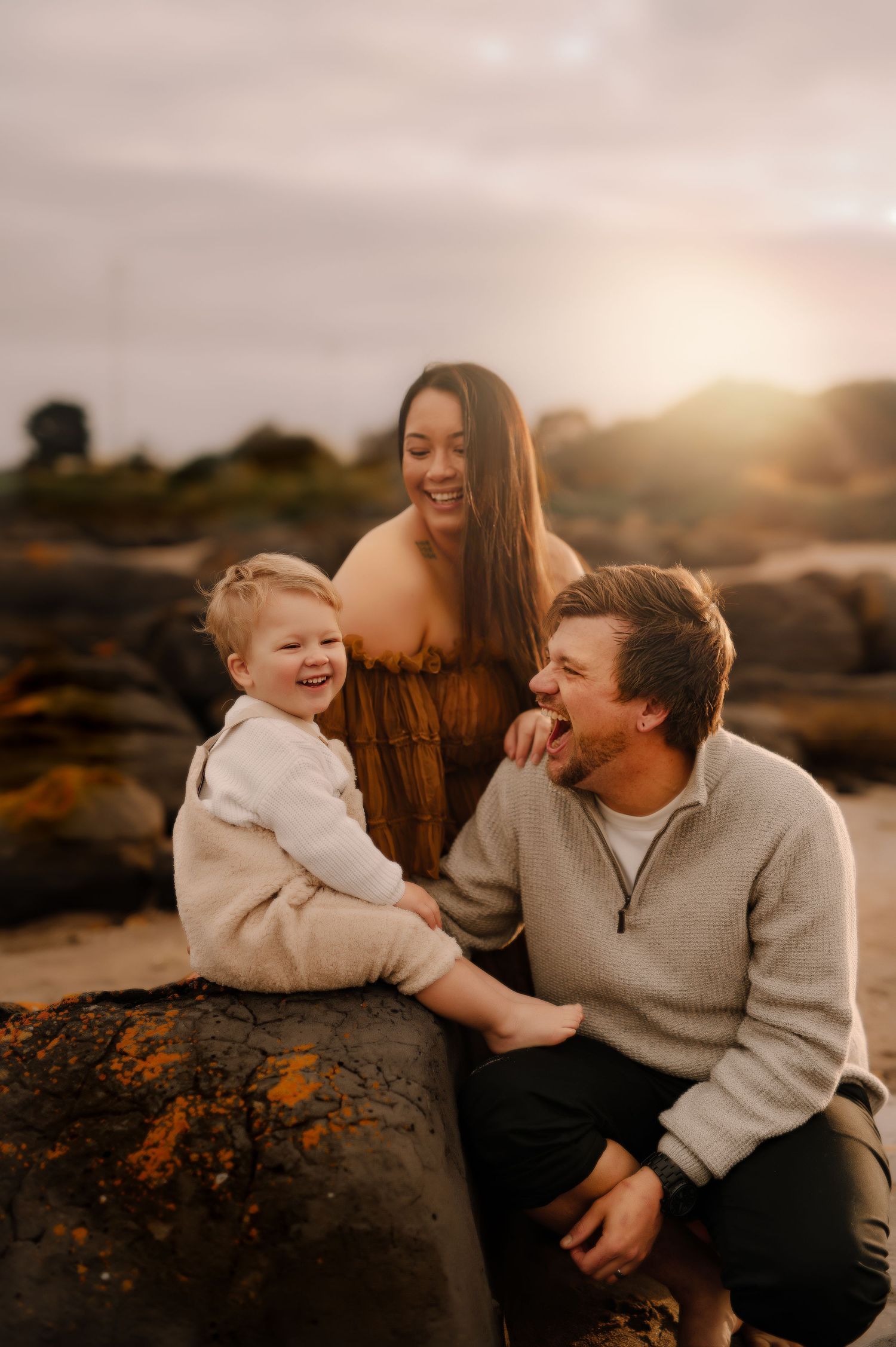 A candid family portrait filled with laughter while sitting on coastal rocks at sunset.