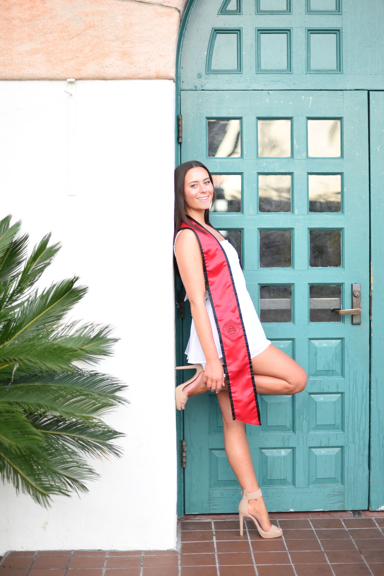 A person in a red and white dress poses gracefully against a turquoise door with palm plants nearby.