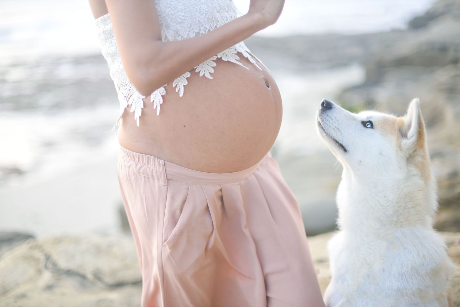 Maternity photos in soft pink and white attire on a sunny beach with natural lighting.