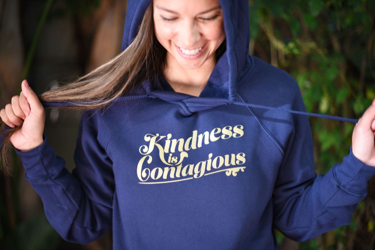 Navy blue hoodie with 'Kindness is Contagious' text being playfully displayed outdoors.
