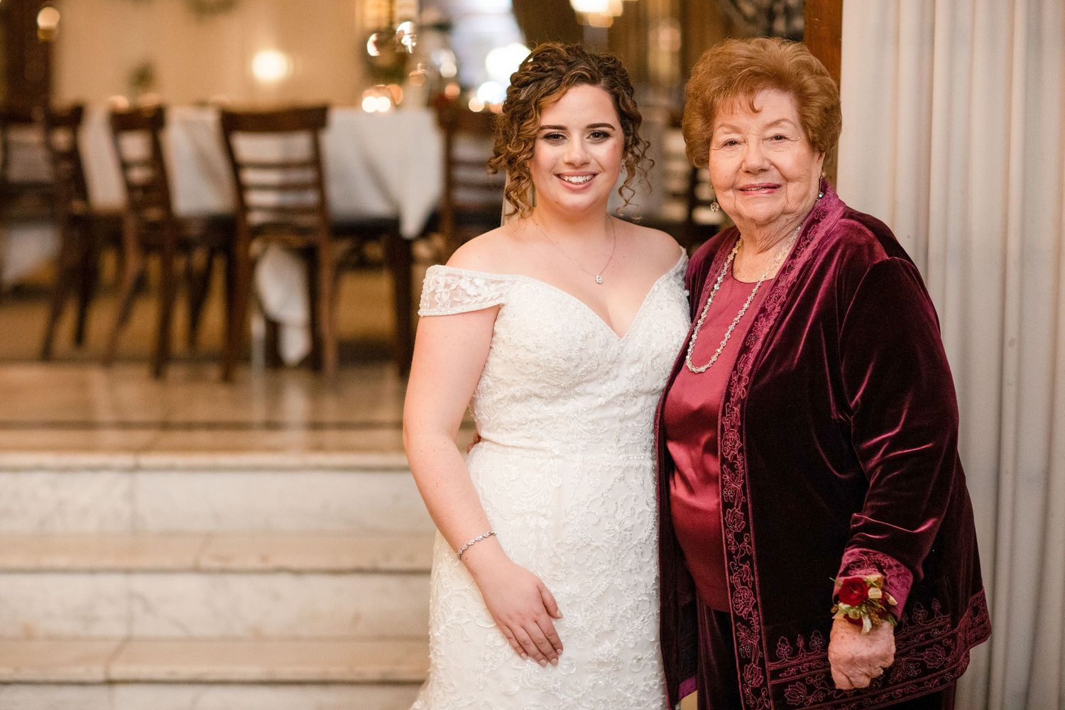 A bride in white dress poses with an older woman wearing a burgundy velvet outfit at a wedding reception.