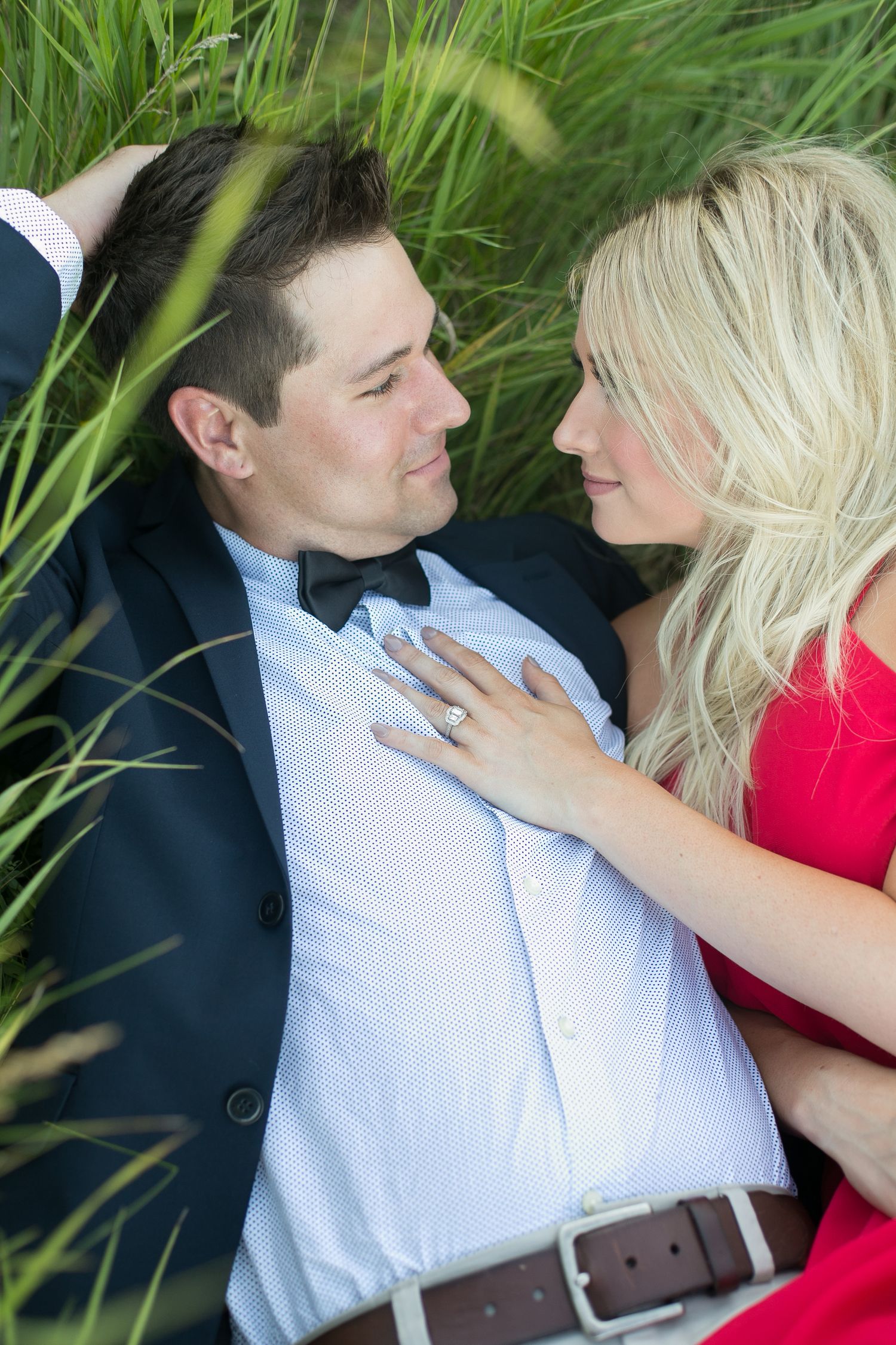 Couple gazing into each others eyes while lying in tall green grass, woman showing engagement ring.