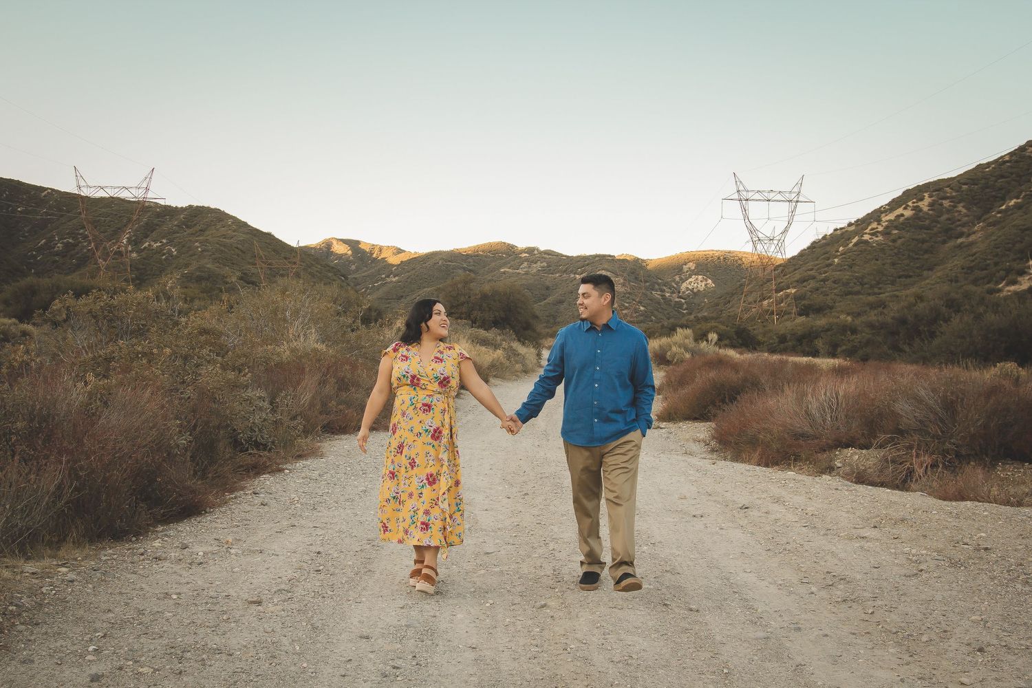 Couple in blue and yellow attire walking hand in hand on a desert trail with mountains in background.