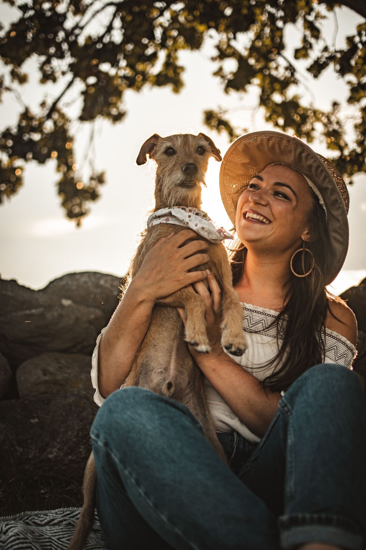 Candid outdoor portrait of woman in hat holding small terrier dog against rocky backdrop.