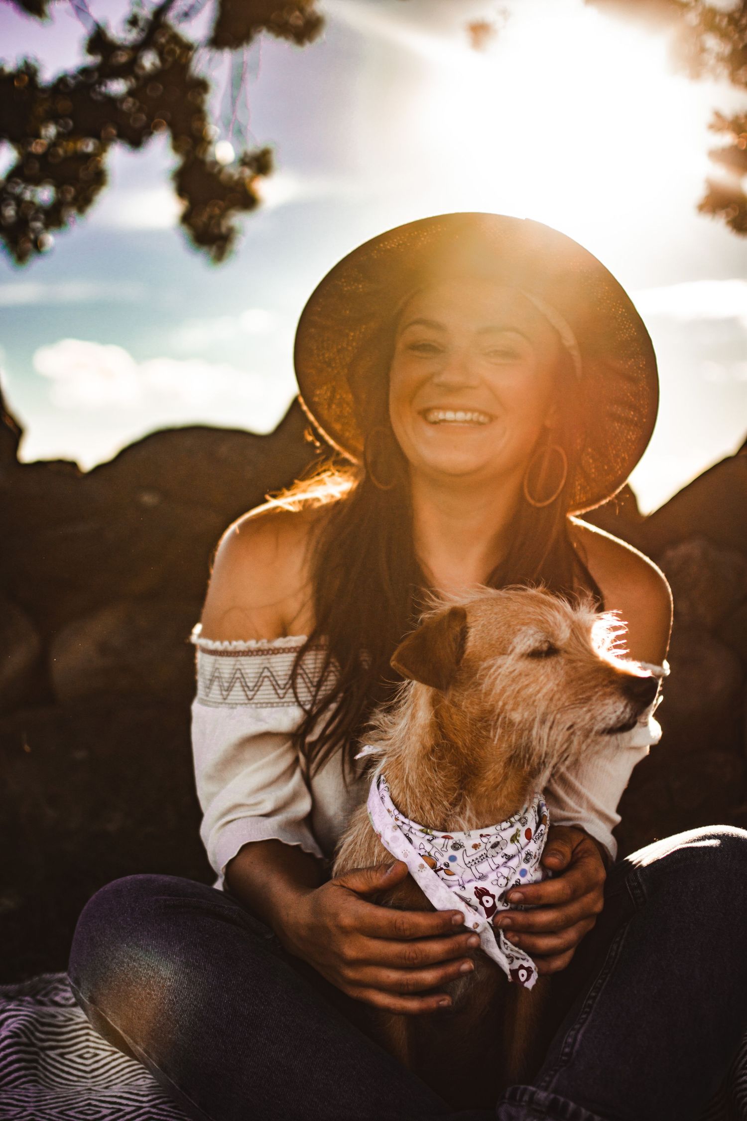 Woman in hat and off-shoulder top sharing joyful moment with dog wearing bandana at sunset.