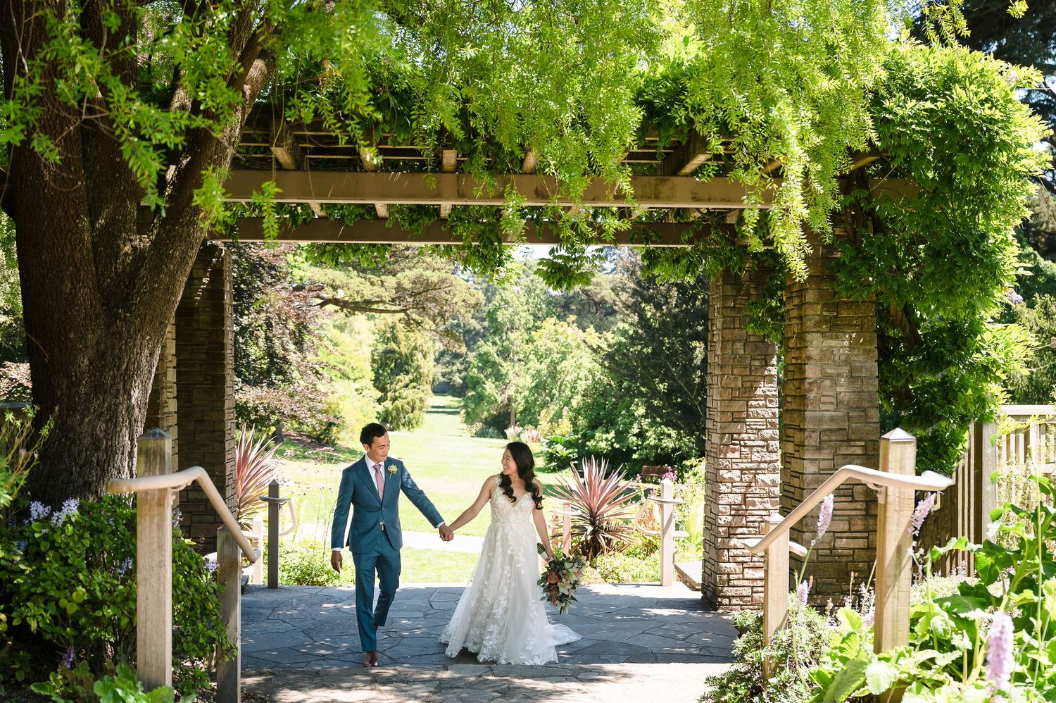 Newlyweds walk hand in hand through a vine-covered brick archway in a garden setting.