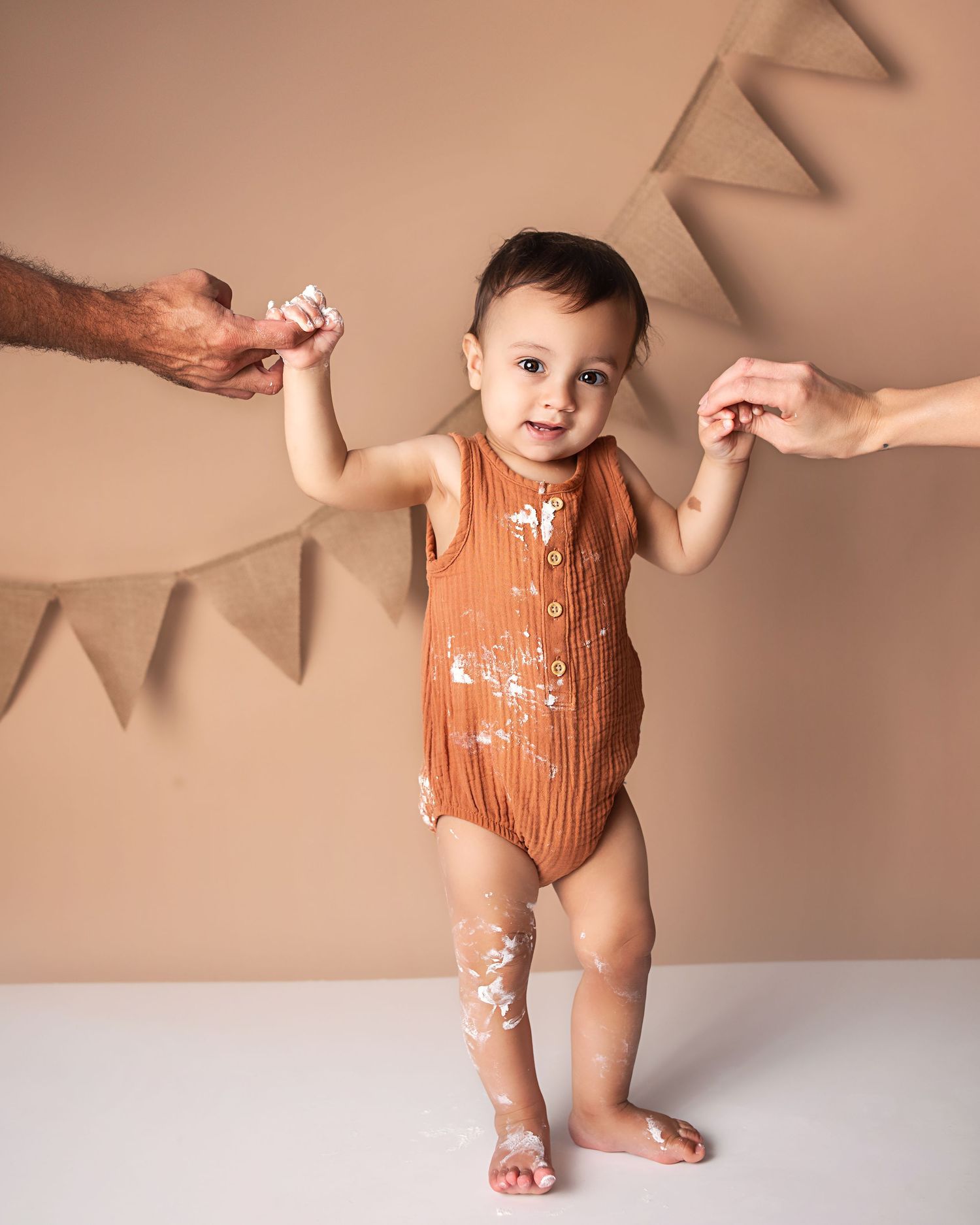 Baby boy in cake-smashed rust romper standing with parent support during first birthday photo session.
