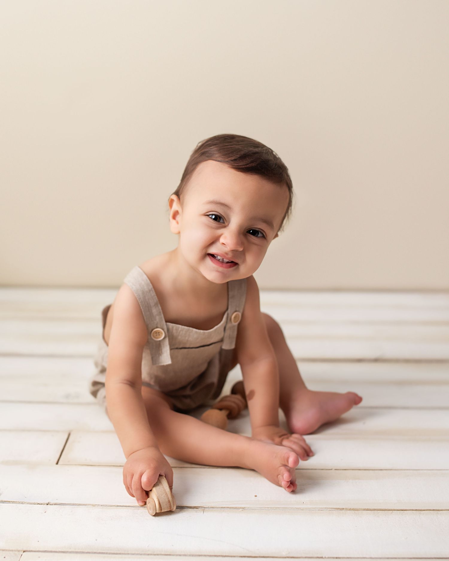 Happy baby boy in linen overalls sitting on white wood floor smiling during milestone photo session.