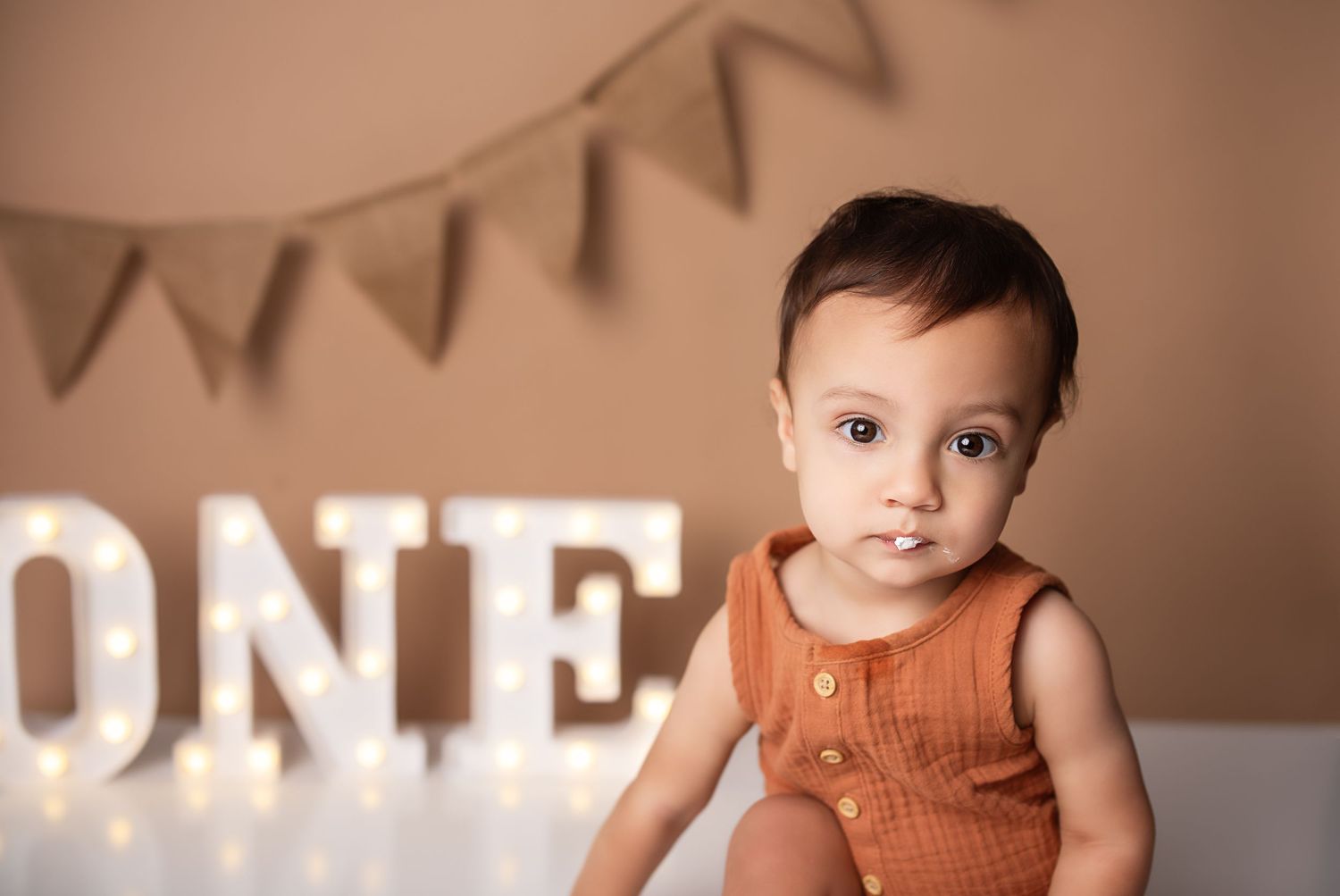 Cute baby boy in rust orange romper posing beside illuminated ONE marquee letters for birthday photos.