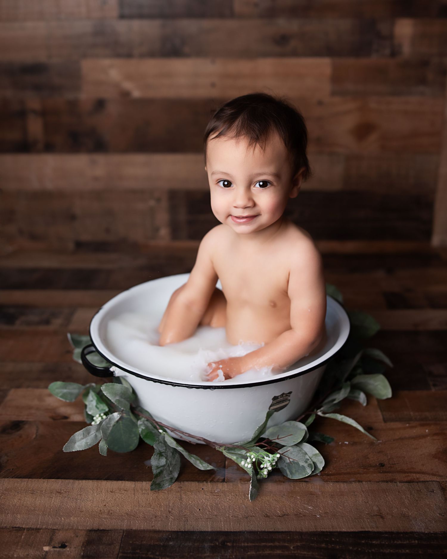 Smiling baby in a white enamel milk bath tub with eucalyptus leaves on rustic wood floor.
