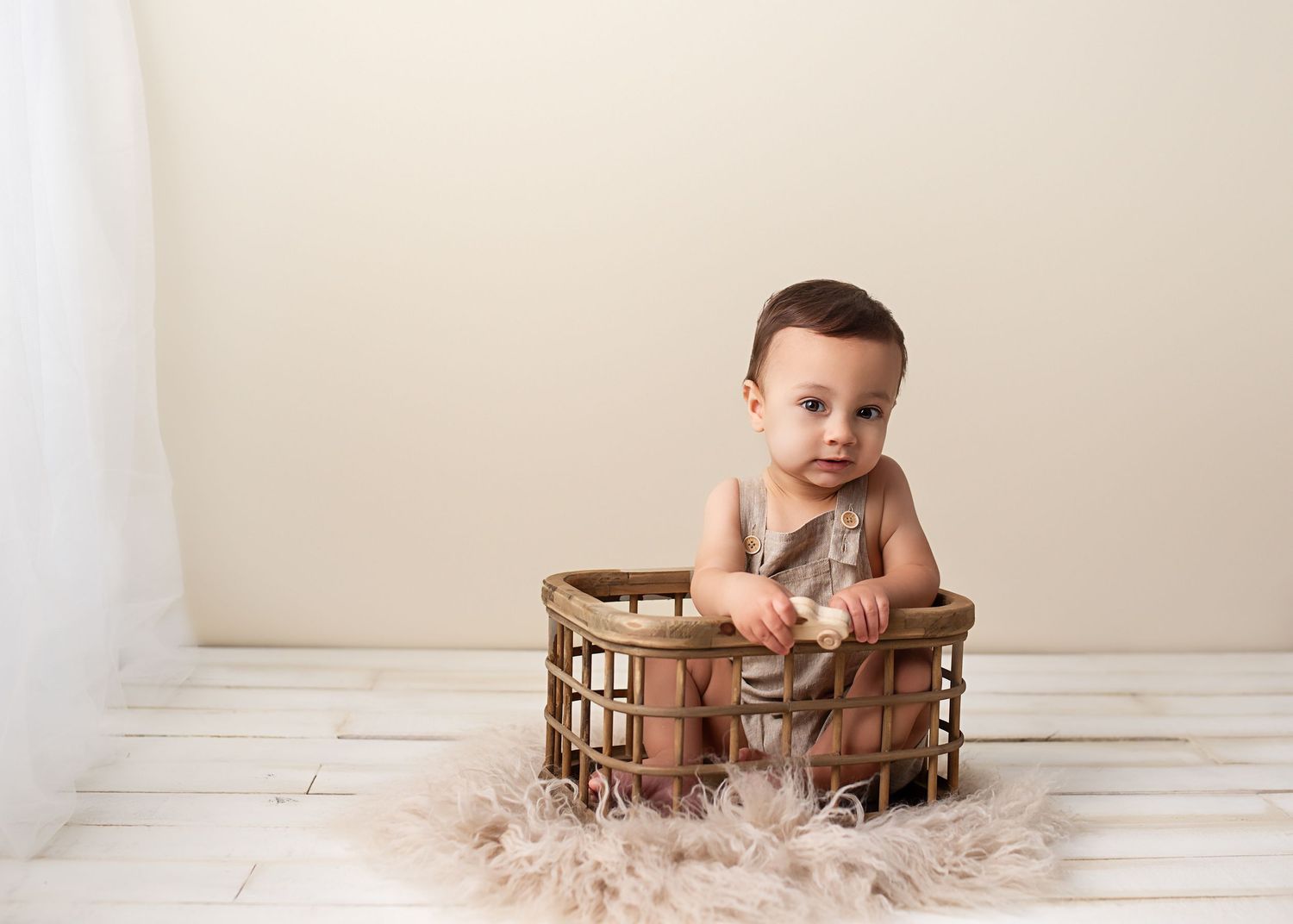 Adorable baby girl in gray romper standing in wicker basket on white wood floor for milestone photos.