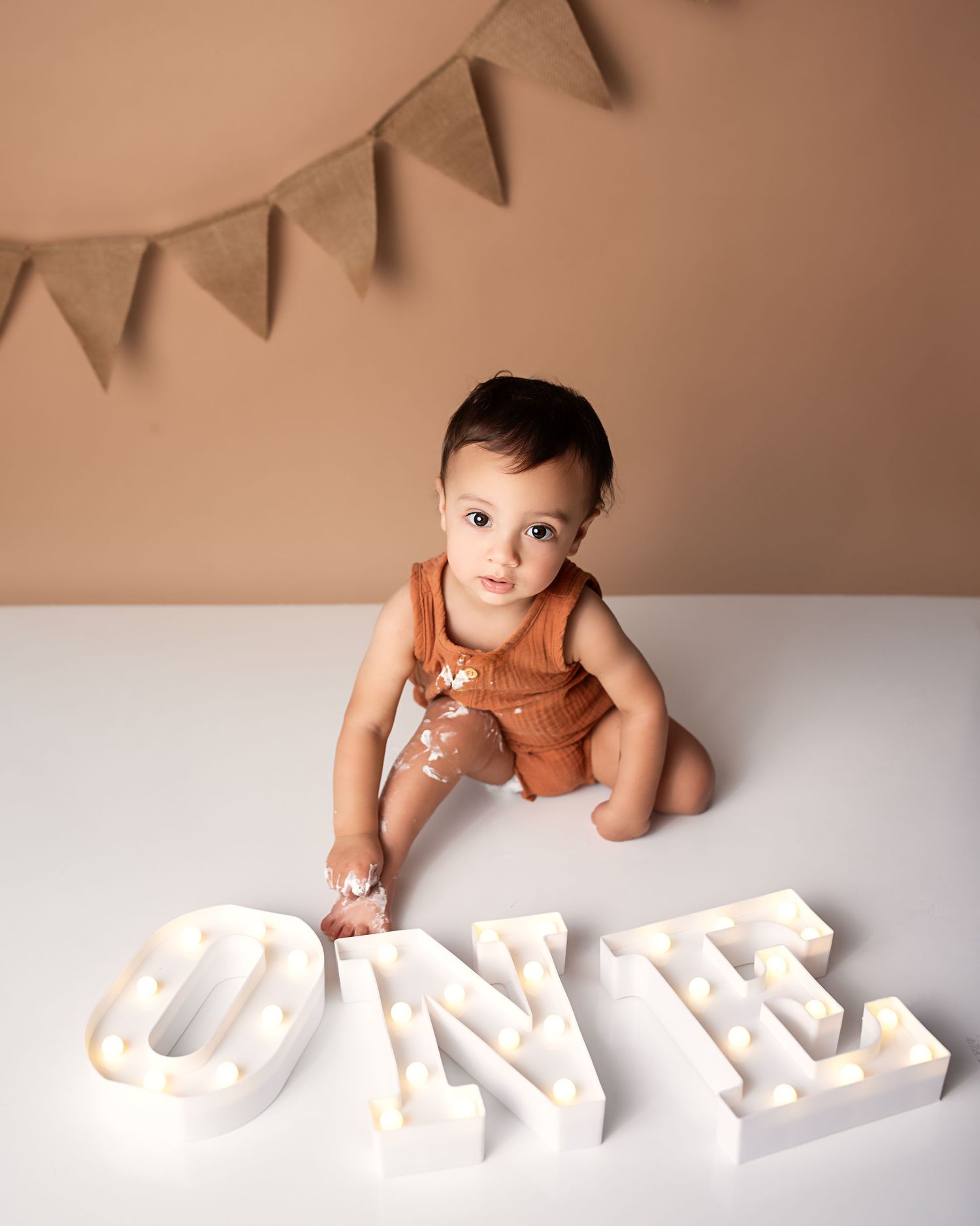 Baby boy in rust romper reaching toward glowing ONE marquee letters during first birthday cake smash.