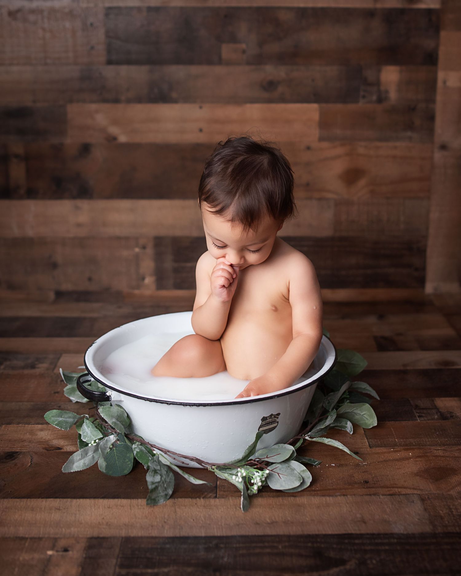 Baby sitting in a white enamel tub with milk bath, surrounded by greenery on rustic wood backdrop.