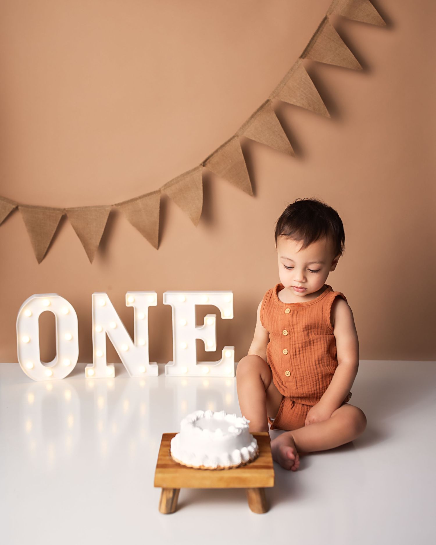 Baby boy in rust romper looking at smash cake beside lit ONE sign for first birthday photo session.