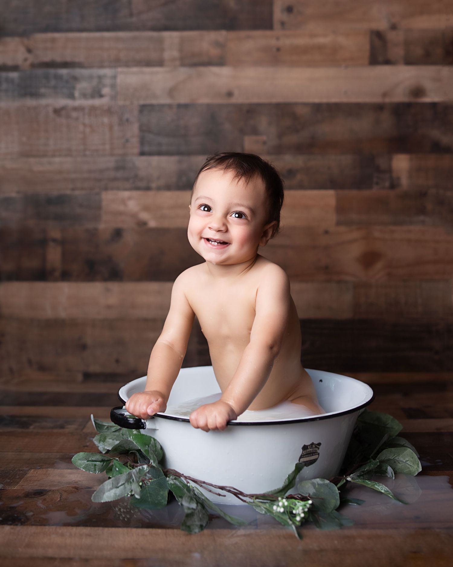 Cheerful baby smiling in a white enamel milk bath tub with greenery on rustic wood floor.