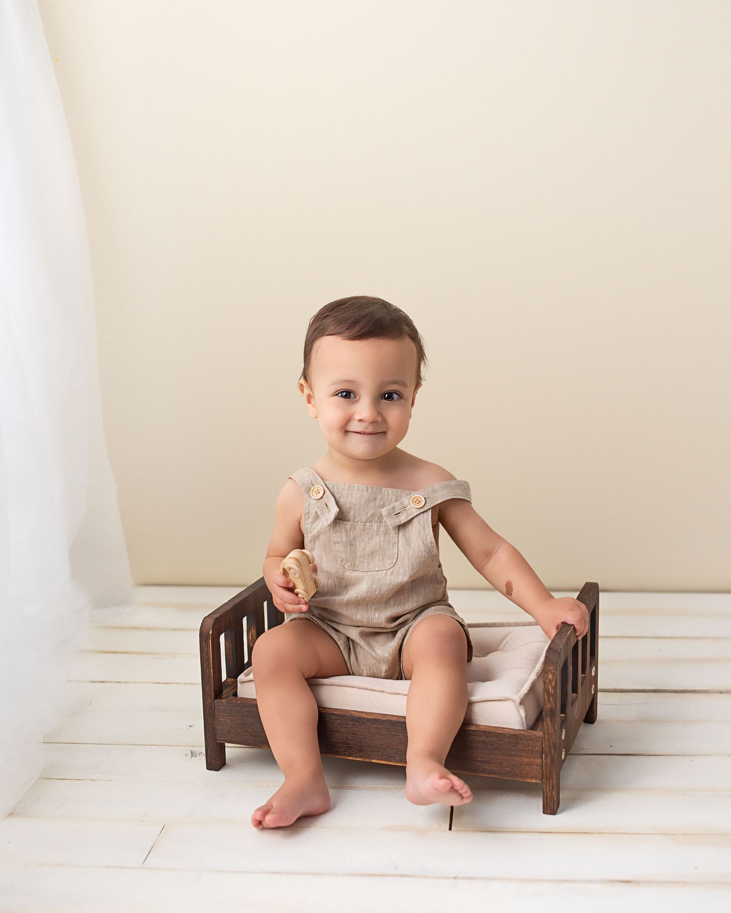 Smiling baby boy in linen romper sitting on miniature wooden bed during professional milestone session.